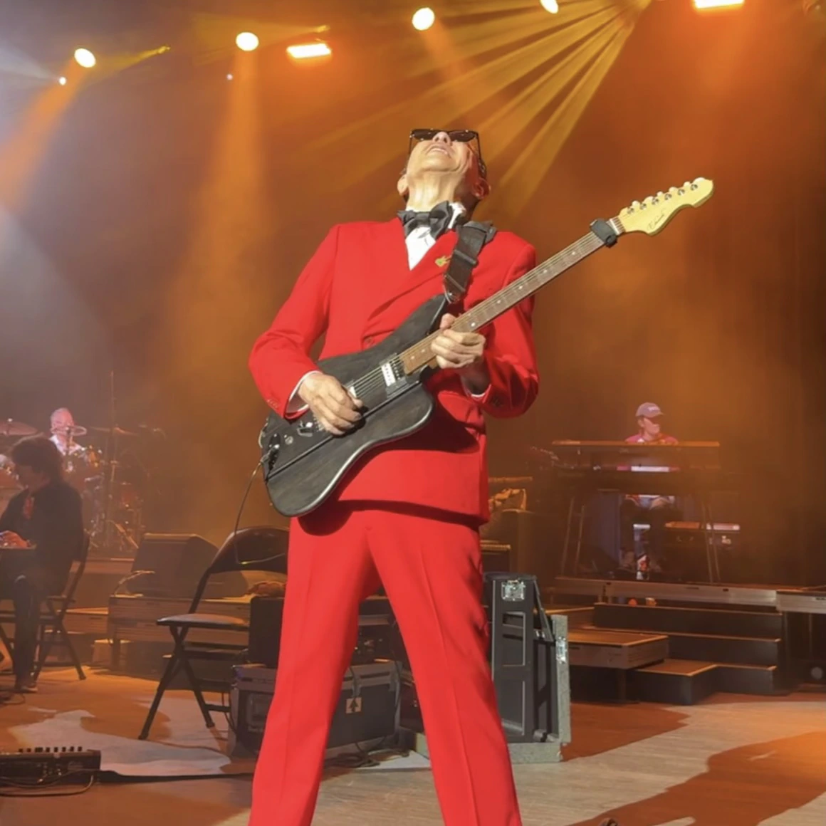 Musician in a red suit playing a black electric guitar under orange stage lighting
