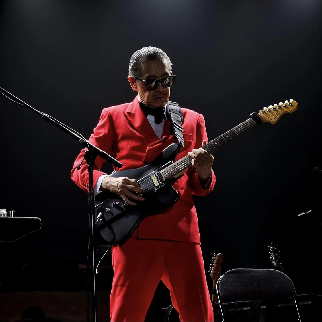 Musician in a red suit playing a black electric guitar under stage lighting