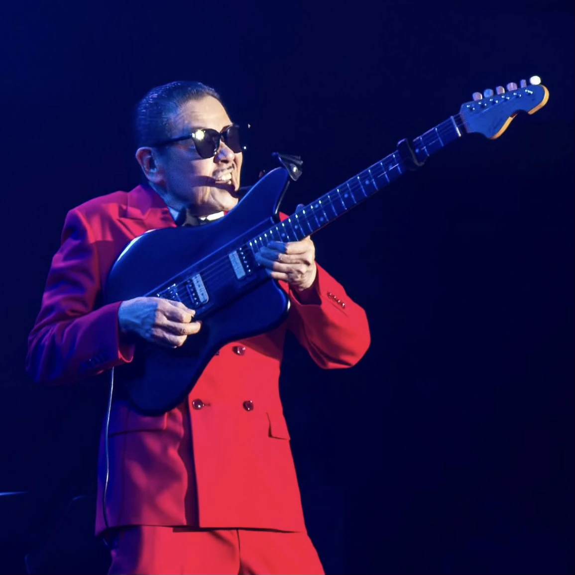 Musician in a red suit playing a black electric guitar under blue stage lighting